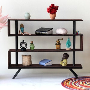 Puede incluir: Una estanter&iacute;a de madera de tres niveles con detalles en metal negro. Las estanter&iacute;as est&aacute;n llenas de varios art&iacute;culos decorativos, incluyendo una mu&ntilde;eca, un libro, una linterna, una botella, un molinillo de caf&eacute;, un &aacute;rbol peque&ntilde;o, un libro, una cesta y una peque&ntilde;a figurita. La estanter&iacute;a est&aacute; sobre una superficie blanca con una alfombra colorida en primer plano.
