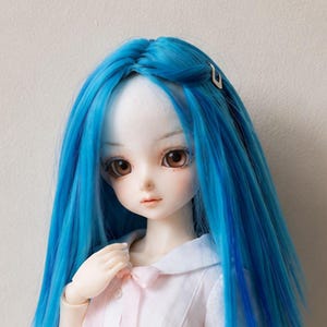 May include: A ball-jointed doll with long, straight, vibrant blue hair featuring a small silver hair clip. The doll has large brown eyes, a pale complexion, and is wearing a white collared shirt with a light pink ribbon tied at the neckline.
