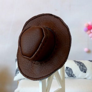 Könnte beinhalten: Ein brauner Cowboyhut aus Leder sitzt auf einem weißen Stuhl in einer Puppenhaus-Szene. Der Hut hat eine breite Krempe und eine spitze Krone.
