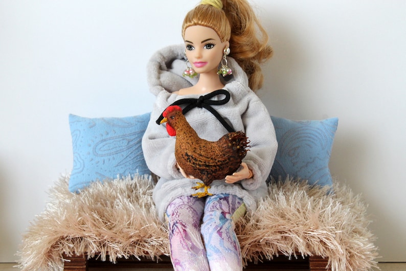 May include: A doll wearing a gray hoodie with a black bow and holding a brown and red toy chicken. The doll is sitting on a light brown fluffy bench with blue pillows.