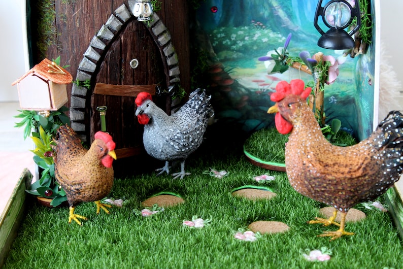 May include: Three miniature chickens in a miniature garden setting. One chicken is gray and white, one is brown and white, and one is brown with black speckles. The chickens are standing on green grass with brown and white flowers.