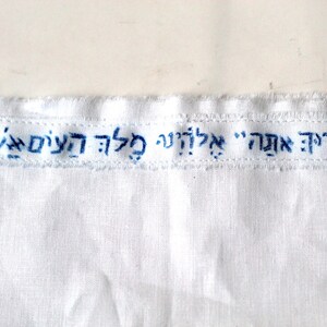 May include: White fabric with blue embroidered text that reads "Fraid Ach" in Hebrew. The text is in a rectangular shape with rounded corners.