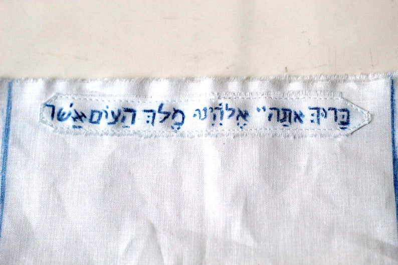 May include: White fabric with blue embroidered text that reads "Fraid Ach" in Hebrew. The text is in a rectangular shape with rounded corners.