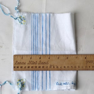 May include: White fabric with blue stripes and two small blue and yellow embroidered patches. A wooden ruler with the text "Senas Mini Land" is placed on top of the fabric.