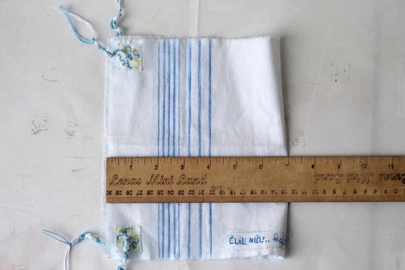 May include: White fabric with blue stripes and two small blue and yellow embroidered patches. A wooden ruler with the text "Senas Mini Land" is placed on top of the fabric.