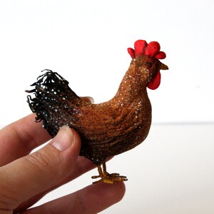 May include: A brown and black chicken figurine with a red comb and wattle. The chicken is standing on a white surface.