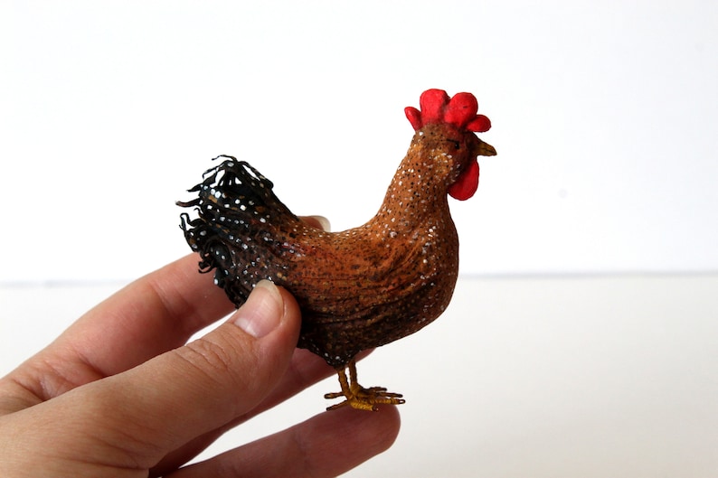 May include: A brown and black chicken figurine with a red comb and wattle. The chicken is standing on a white surface.