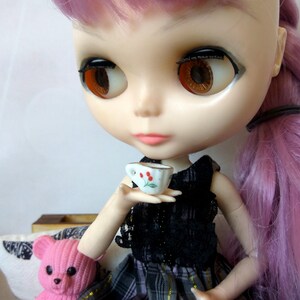 May include: A doll with long pink hair is holding a small white teacup with a red flower design. The doll is wearing a black and white dress with a plaid skirt.