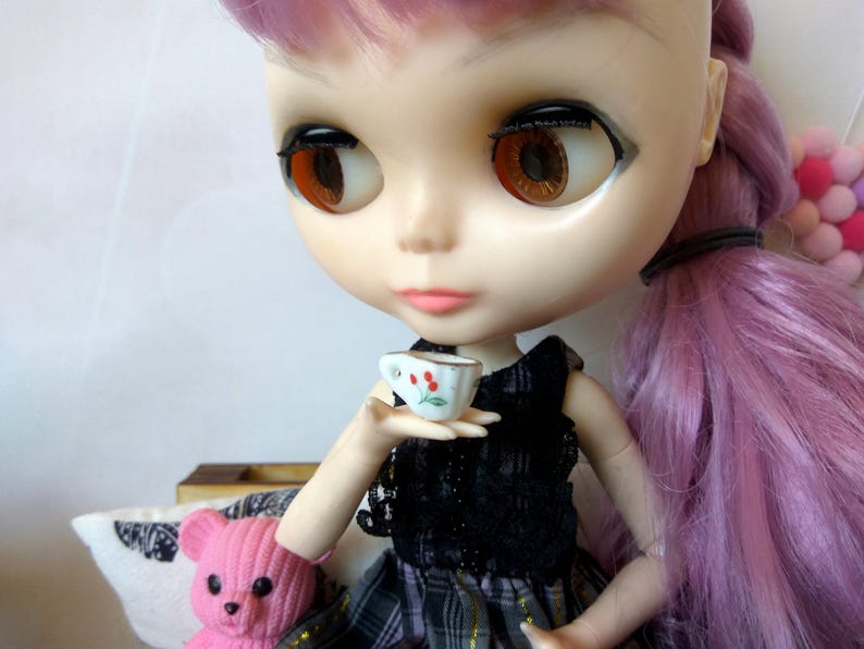 May include: A doll with long pink hair is holding a small white teacup with a red flower design. The doll is wearing a black and white dress with a plaid skirt.