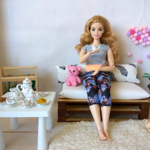 May include: A doll sitting on a miniature couch with a pink teddy bear, a tea set on a white table, and a pink pom-pom heart hanging on the wall.