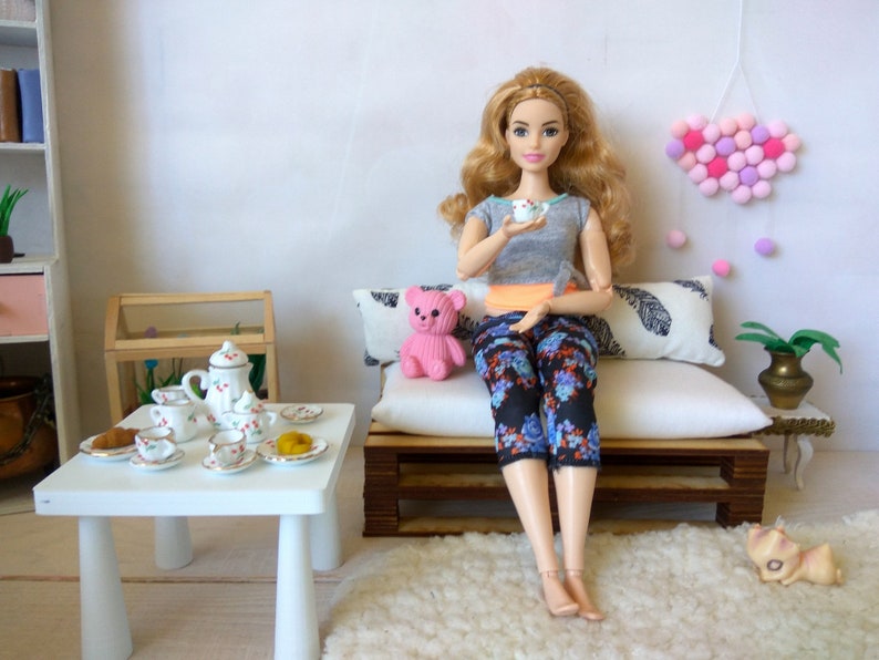 May include: A doll sitting on a miniature couch with a pink teddy bear, a tea set on a white table, and a pink pom-pom heart hanging on the wall.