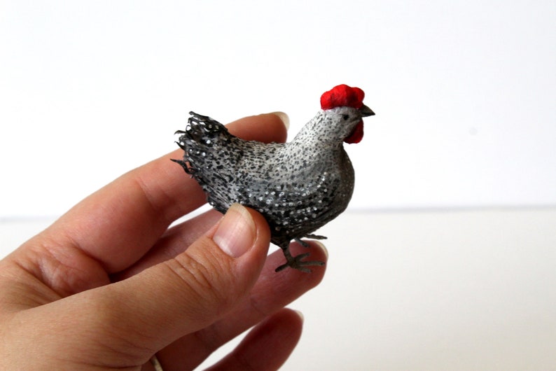 May include: A small, gray and white chicken figurine with a red comb. The chicken is standing on a hand.