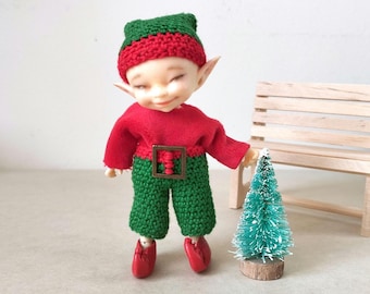 May include: A small doll with a light skin tone, wearing a green and red crocheted hat, a red long-sleeved top, and green crocheted pants with a brown square belt buckle. The doll has red shoes and stands next to a miniature green bottle brush tree.