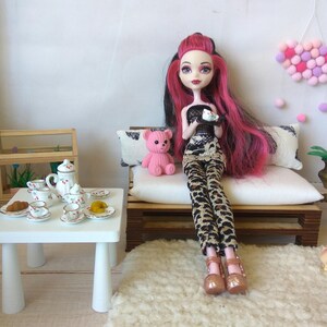 May include: A doll with pink hair and a black and white outfit sits on a white couch with a pink teddy bear. The doll is holding a small white plate with a pink and white dessert. The couch is in a dollhouse setting with a white table and tea set.