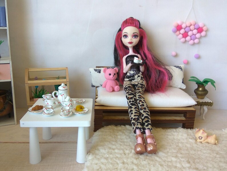 May include: A doll with pink hair and a black and white outfit sits on a white couch with a pink teddy bear. The doll is holding a small white plate with a pink and white dessert. The couch is in a dollhouse setting with a white table and tea set.
