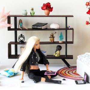 Puede incluir: Una mu&ntilde;eca sentada en el suelo con una tableta rosa en su regazo. La mu&ntilde;eca lleva una chaqueta de cuero negra, una camisa a rayas, pantalones negros y botas negras. La mu&ntilde;eca est&aacute; sentada frente a una estanter&iacute;a con varios art&iacute;culos en los estantes, incluyendo un libro, un jarr&oacute;n, una linterna, un molinillo de caf&eacute; y un peque&ntilde;o &aacute;rbol de Navidad. Hay un otomano blanco con un coj&iacute;n estampado en el lado derecho de la imagen.