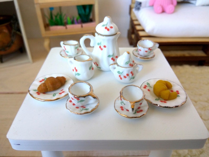 May include: A white porcelain tea set with a cherry design. The set includes a teapot, creamer, sugar bowl, two cups and saucers, and two plates with croissants.