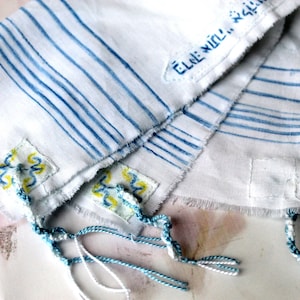 Peut inclure: Tissu blanc à rayures bleues et broderie jaune et bleue. Le tissu est plié en deux et a des glands en bas. Le texte "Elle Will..." est visible sur le tissu.