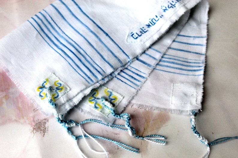 May include: White fabric with blue stripes and yellow and blue embroidery. The fabric is folded in half and has tassels on the bottom. The text "Elle Will..." is visible on the fabric.