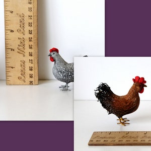 May include: Two handmade felt chickens, one brown with a red comb and one gray with a red comb. The chickens are standing on a white surface next to a wooden ruler.