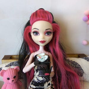 May include: A pink haired doll with black streaks in her hair is sitting on a couch. She is wearing a black and white dress and holding a small white teacup with a floral design.