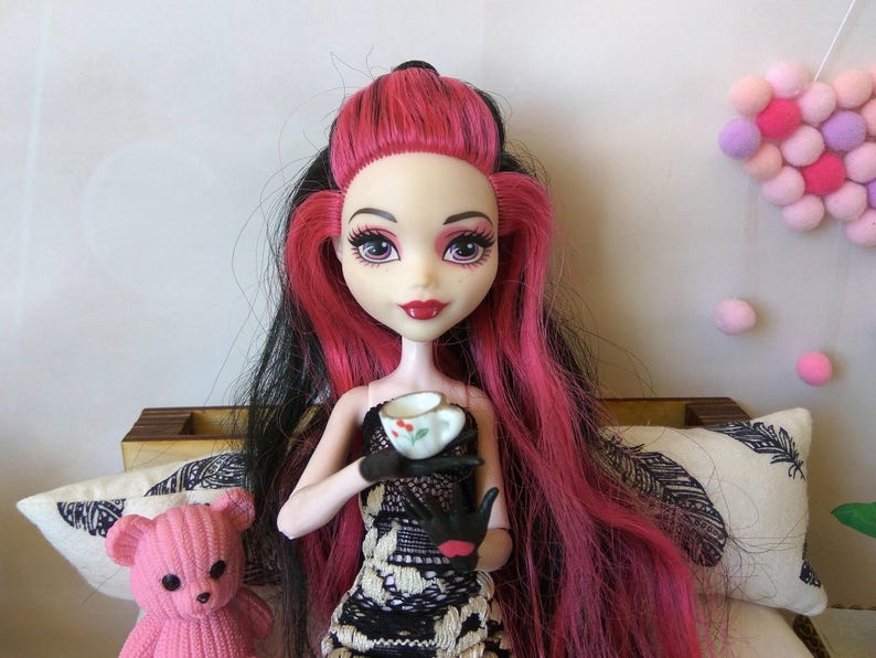May include: A pink haired doll with black streaks in her hair is sitting on a couch. She is wearing a black and white dress and holding a small white teacup with a floral design.