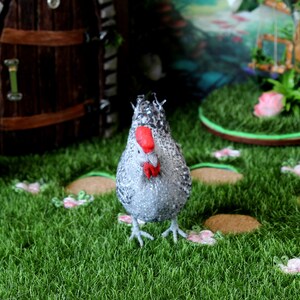 May include: A gray and white chicken figurine with a red comb and wattle stands on a green artificial grass surface. The figurine is surrounded by small brown circles and pink flowers.
