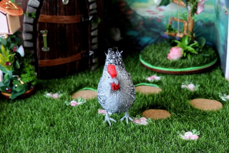 May include: A gray and white chicken figurine with a red comb and wattle stands on a green artificial grass surface. The figurine is surrounded by small brown circles and pink flowers.
