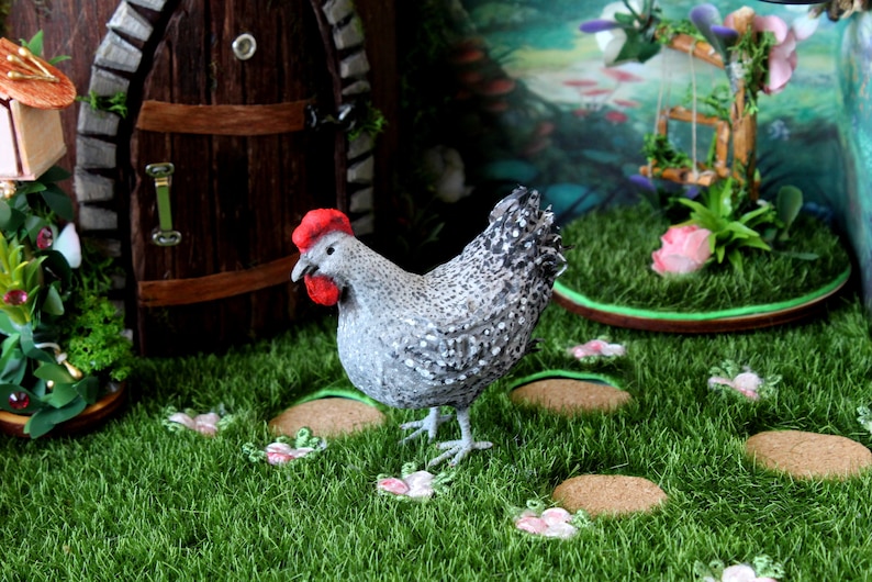 May include: A gray and white speckled chicken figurine with a red comb stands on a green grassy surface with small pink flowers. The chicken is facing the left side of the image.