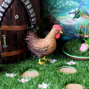 May include: A miniature fairy garden scene with a brown wooden door, a small mailbox, a brown and white chicken figurine, and a swing set with green vines. The garden is decorated with green grass, pink flowers, and a green and brown circular base.