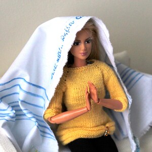 May include: A doll wearing a yellow sweater and a white headscarf with blue stripes and the text "The Win..." printed on it. The doll is sitting on a white chair with a blue and white striped throw.