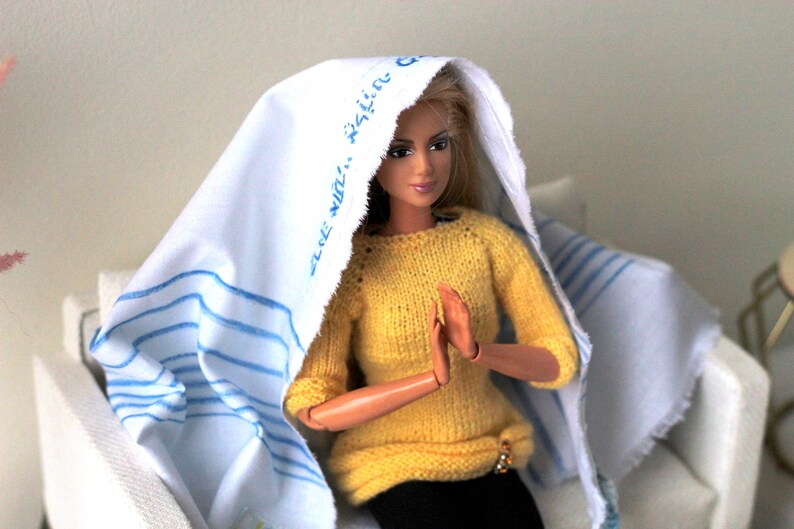 May include: A doll wearing a yellow sweater and a white headscarf with blue stripes and the text "The Win..." printed on it. The doll is sitting on a white chair with a blue and white striped throw.
