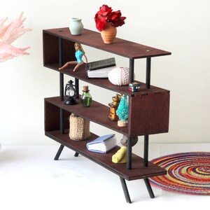 Puede incluir: Una estanter&iacute;a de madera de tres niveles con patas y soportes de metal negro. Las estanter&iacute;as est&aacute;n llenas de art&iacute;culos decorativos, incluyendo una mu&ntilde;eca, un libro, una concha, una linterna, una botella, una cesta, un &aacute;rbol peque&ntilde;o y un libro. La estanter&iacute;a est&aacute; sobre una superficie blanca con una alfombra colorida en primer plano.
