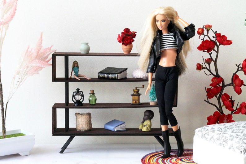 Puede incluir: Una estanter&iacute;a de metal negro con tres estantes, que presenta una variedad de art&iacute;culos decorativos, incluyendo una mu&ntilde;eca, un libro, una linterna, un jarr&oacute;n, una planta y un molinillo de caf&eacute;. La estanter&iacute;a est&aacute; contra una pared blanca.