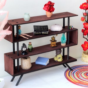 Puede incluir: Una estanter&iacute;a de madera marr&oacute;n con patas de metal negro y tres estantes. Los estantes est&aacute;n decorados con varios art&iacute;culos, incluyendo un libro, una mu&ntilde;eca, una linterna, una botella, un molinillo de caf&eacute;, un &aacute;rbol peque&ntilde;o, un erizo de mar, una peque&ntilde;a estatua y un libro azul. La estanter&iacute;a est&aacute; sobre una superficie blanca.