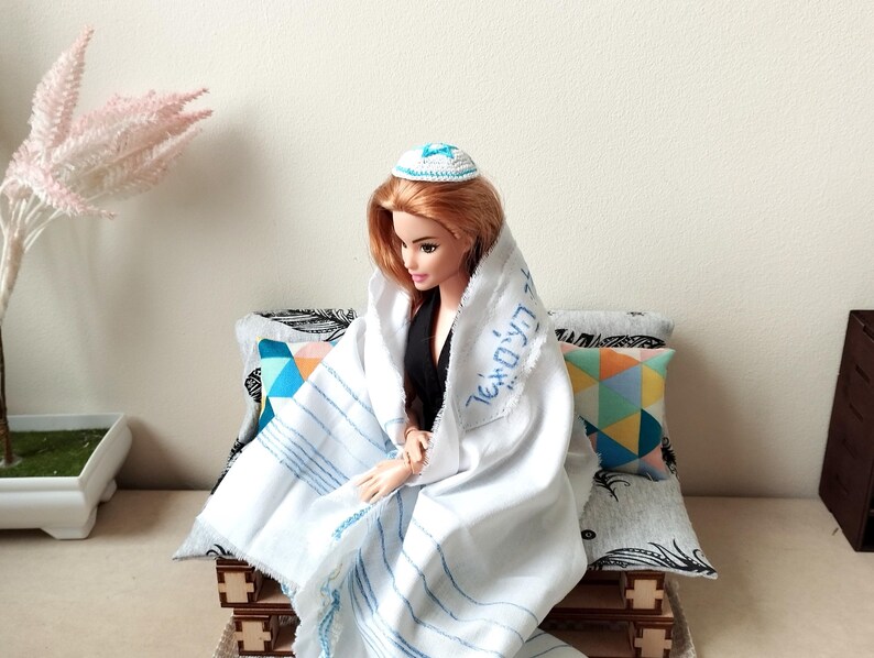 May include: A doll wearing a white prayer shawl with blue stripes and a blue and white kippah. The doll is sitting on a couch with colorful pillows.