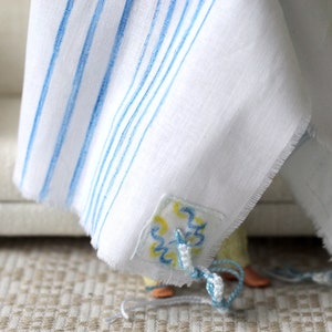 May include: A white prayer shawl with blue stripes and a decorative blue and yellow embroidered patch. The shawl is draped over a chair.