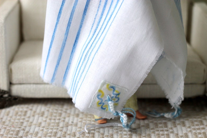May include: A white prayer shawl with blue stripes and a decorative blue and yellow embroidered patch. The shawl is draped over a chair.