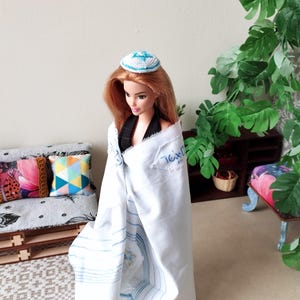 May include: A doll wearing a white prayer shawl with blue stripes and a blue and white kippah. The doll is standing in front of a couch with colorful pillows and a plant in the background.