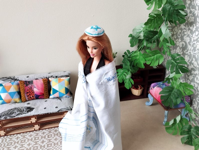May include: A doll wearing a white prayer shawl with blue stripes and a blue and white kippah. The doll is standing in front of a couch with colorful pillows and a plant in the background.