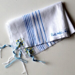 May include: White prayer shawl with blue stripes and blue and yellow floral embroidery. The shawl has fringes and the text "&Eacute;LE N&Iacute;LL..." is written on it.