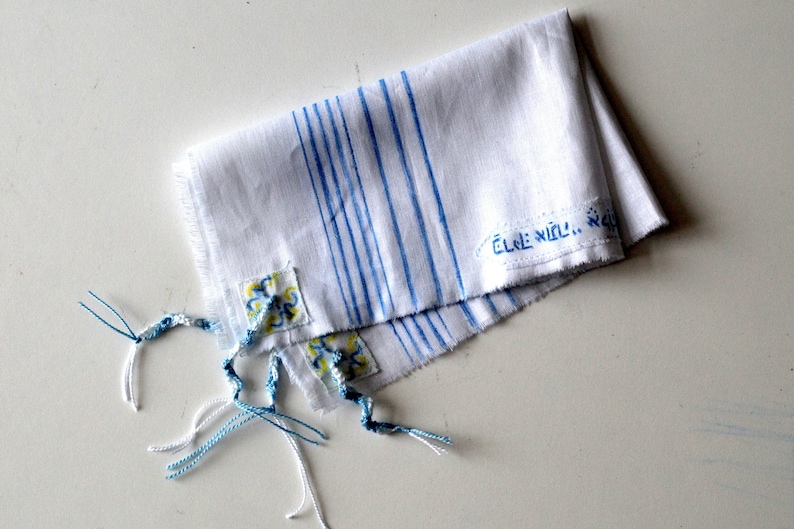 May include: White prayer shawl with blue stripes and blue and yellow floral embroidery. The shawl has fringes and the text "&Eacute;LE N&Iacute;LL..." is written on it.