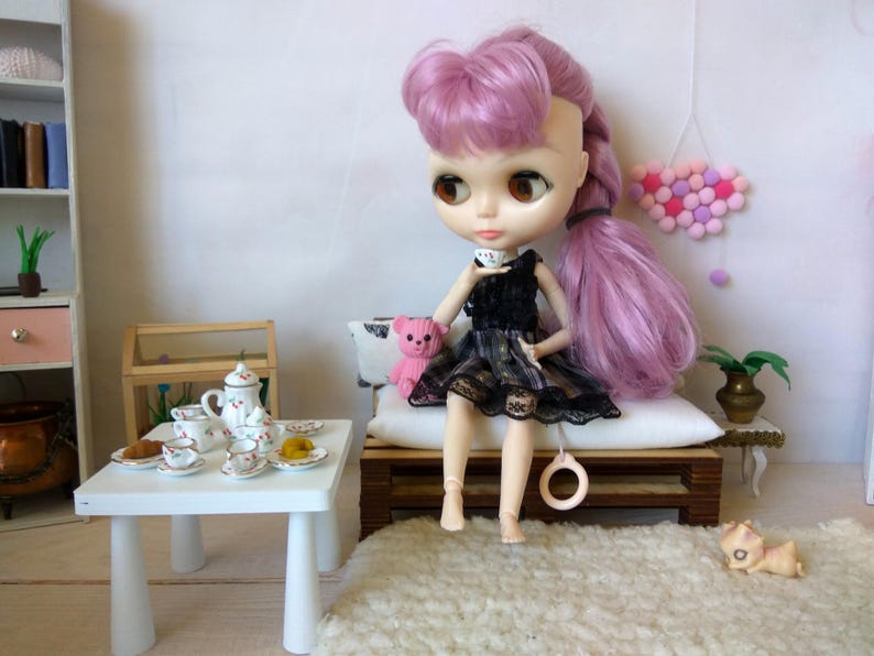 May include: A doll with pink hair and a black dress sits on a wooden bench with a pink teddy bear. The doll is holding a teacup and there is a white table with a tea set on it.