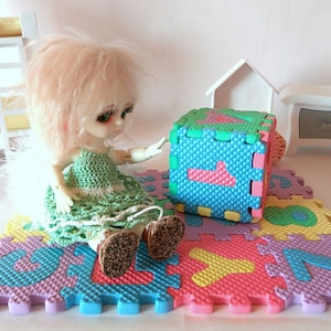May include: A doll with blonde hair wearing a green crocheted dress plays with a colorful foam puzzle mat. The mat features numbers and is perfect for a child's playroom.