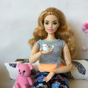 May include: A doll with long blonde hair is sitting on a couch with a pink teddy bear. She is holding a small white teacup with red flowers on it.