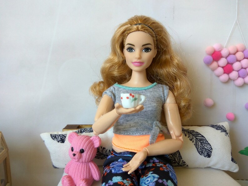 May include: A doll with long blonde hair is sitting on a couch with a pink teddy bear. She is holding a small white teacup with red flowers on it.