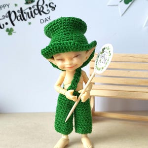 May include: A small doll wearing a green crocheted hat and overalls, holding a sign that says "Happy St. Patrick's Day". The doll is sitting on a wooden bench.