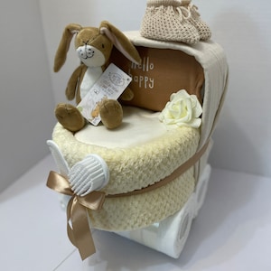 May include: A white baby carriage diaper cake with a brown bunny plush toy, a pair of knitted booties, a white rose, and a brown and white tag that says "Hello Happy".