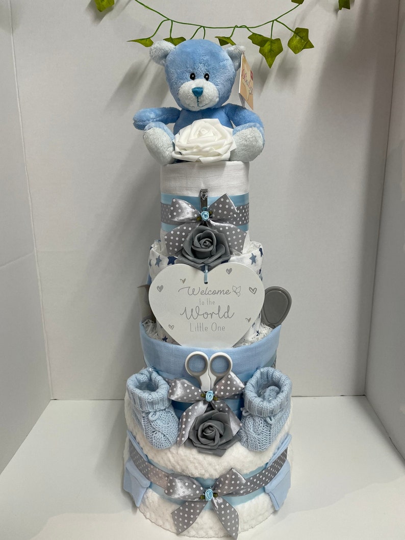 A tall 4 tier nappy cake in blue with nappies, baby blanket, 3 x muslin squares of different shades of blue, baby booties, brush set, bib, hat, scratch mitts, manicure set and a soft blue teddy bear. Wrapped ready to gift including ribbons and tag