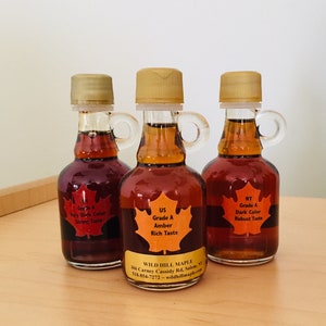 May include: Three small glass bottles of maple syrup with gold-colored caps and handles. Each bottle has a label with a maple leaf design and text indicating the grade and taste: Very Dark, Amber, and Dark. The bottles are on a light-colored wooden surface.
